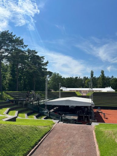 Blick auf eine Bühne in einem Freiluft-Ampitheater mit Bäumen und blauem Himmel.