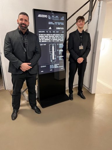 Zwei Männer in formeller Kleidung stehen neben einem Informationsdisplay.